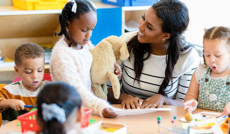 The mid adult female teacher manages the classroom full of busy young children.