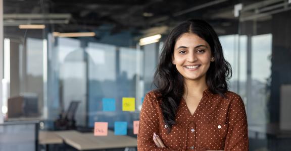 confident businesswoman at workplace inside office, woman with crossed arms smiling and looking at camera, standing near window