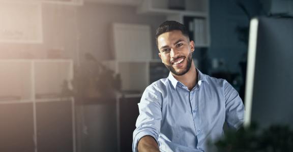 young man smiling at a camera while he uses his computer,in his office
