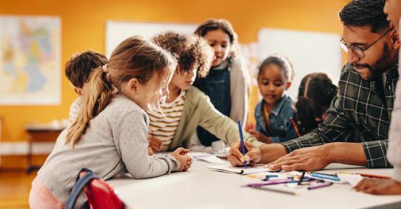 Teacher showing students how to draw using a color pencil around a table