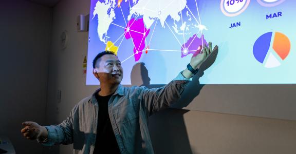 University lecturer pointing at a projector screen with an open palm.