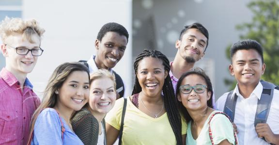 A group of eight multi-ethnic teenagers, 17 and 18 years old, carrying book bags, standing together outside a school building.