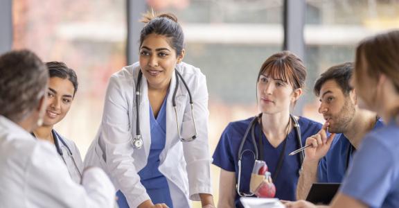 A small medical team of professionals sit around a boardroom table as they meet together to discuss patient cases. T