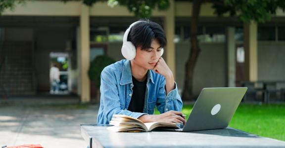 Young Asian student working at a laptop outside at a picnic table.