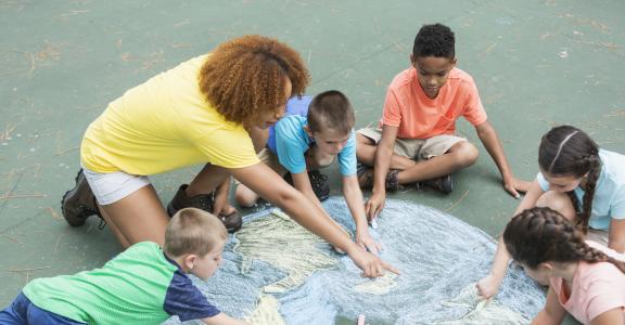 Teacher with five multi-ethnic children at summer camp, sitting on a playground, working together to make a large chalk drawing of planet earth.