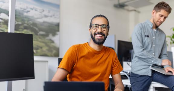 happy young man sitting at desk with colleague in background at office. 