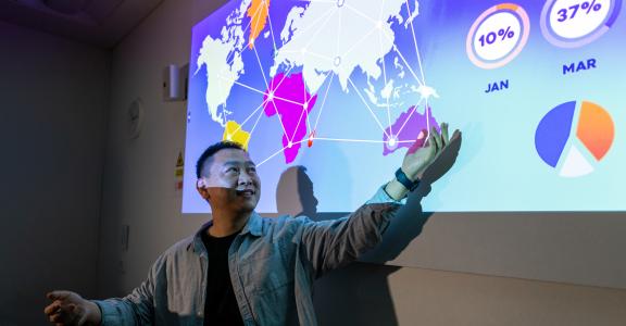University lecturer pointing at a projector screen with an open palm. The projector screen shows a presentation about data mining.