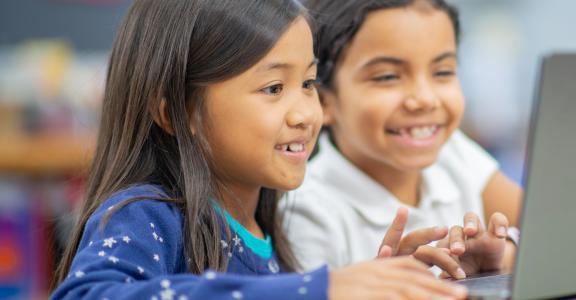 Two enthusiastic children are collaborating on laptops in a bright classroom setting, exhibiting teamwork and engagement while participating in a technology-based educational activity.