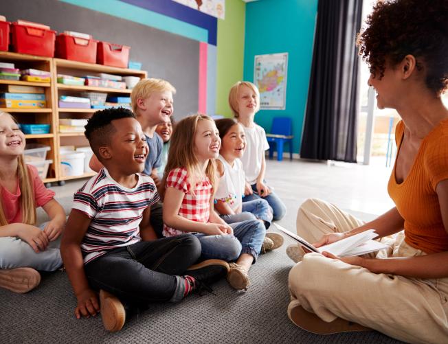 Group of elementary school students sitting on floor listening to female teacher read book