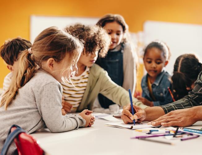 Teacher showing students how to draw using a color pencil around a table