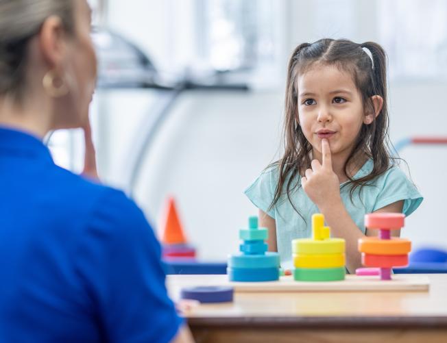Female speech therapist sitting at a table with a young girl, pointing to her mouth to demonstrate word formation