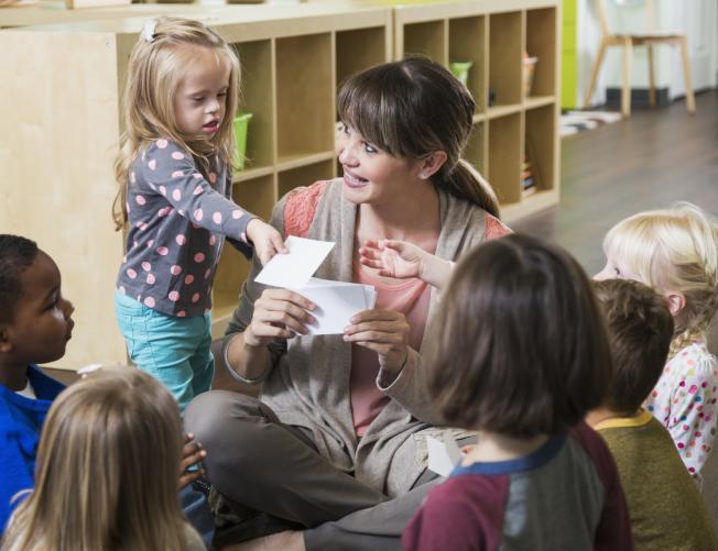 Preschool teacher sitting with multi-ethnic children in a circle; a girl with special needs hands a card to another child