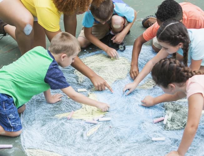 Instructor with five multi-ethnic children at summer camp, sitting on a playground, working together to make a large chalk drawing of planet earth.