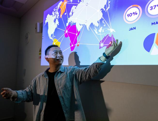 University lecturer pointing at a projector screen with an open palm. The projector screen shows a presentation about data mining.