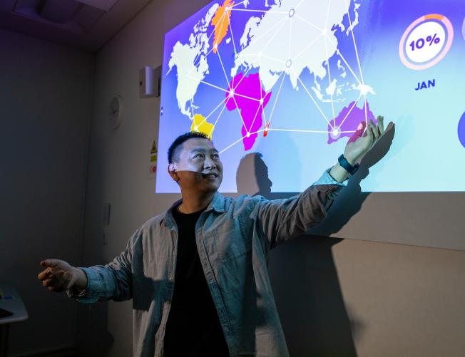 University lecturer pointing at a projector screen with an open palm. The projector screen shows a presentation about data mining.