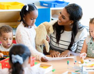 The mid adult female teacher manages the classroom full of busy young children.
