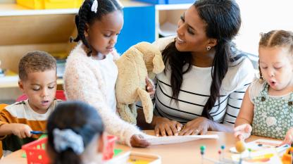 The mid adult female teacher manages the classroom full of busy young children.