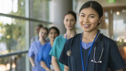 A female doctor of Asian descent poses with her multi-ethnic team of doctors and nurses and smiles directly at the camera while standing in a hospital corridor.