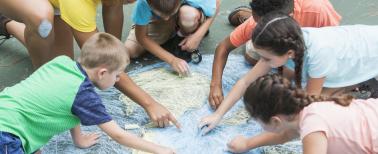 Instructor with five multi-ethnic children at summer camp, sitting on a playground, working together to make a large chalk drawing of planet earth.