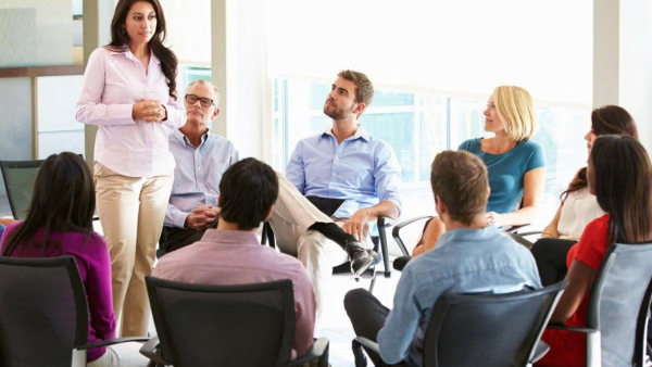 Group of people sitting in a circle looking up at a female speaker. 