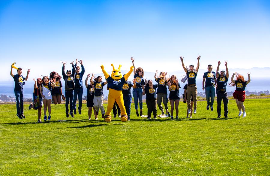 A group of students jumping in a line on a grassy field with Sammy the mascot